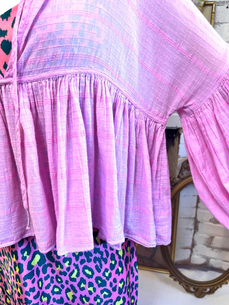 May include: A pink blouse with a ruffled hem and long, flared sleeves. The top has a textured, woven appearance and a drawstring neckline. The lower portion of the image shows a skirt with a leopard print design in pink, green, and black.