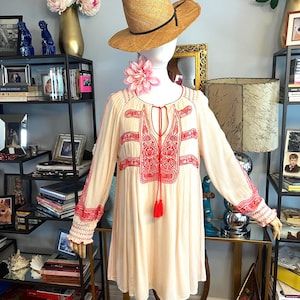 May include: A cream tunic dress with red embroidery and tassel details. The dress has long sleeves with embroidered cuffs. A straw hat with a black band sits on a mannequin head, with a pink flower necklace. The dress is displayed on a glass table.