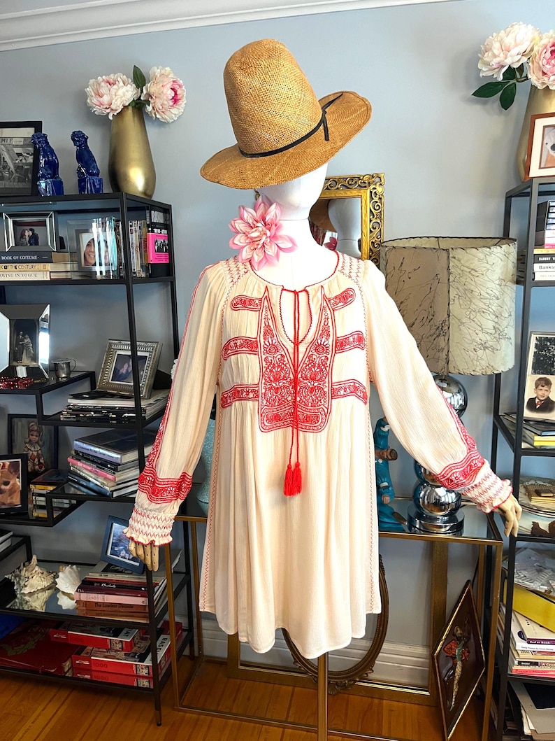 May include: A cream tunic dress with red embroidery and tassel details. The dress has long sleeves with embroidered cuffs. A straw hat with a black band sits on a mannequin head, with a pink flower necklace. The dress is displayed on a glass table.