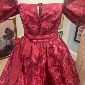 May include: A vibrant, short, red dress with a floral pattern. The dress features a square neckline, puffed sleeves, and a tied detail at the bust. A matching belt with a large bow accentuates the waist. The dress is displayed on a mannequin.