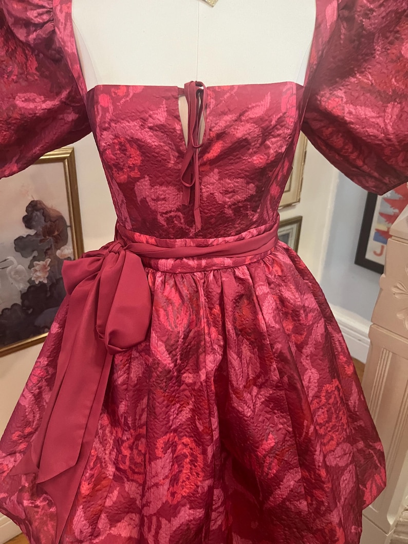 May include: A vibrant, short, red dress with a floral pattern. The dress features a square neckline, puffed sleeves, and a tied detail at the bust. A matching belt with a large bow accentuates the waist. The dress is displayed on a mannequin.