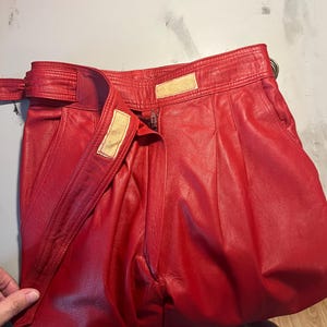 May include: Bright red leather pants with a high waist and a belt. The pants have a zipper closure and a hook-and-loop fastener. The leather appears soft and has a slight sheen. The pants are folded, showing the pleats and the belt detail.