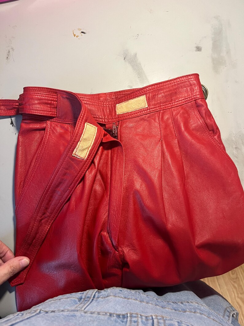 May include: Bright red leather pants with a high waist and a belt. The pants have a zipper closure and a hook-and-loop fastener. The leather appears soft and has a slight sheen. The pants are folded, showing the pleats and the belt detail.