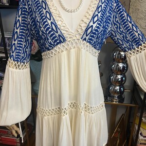 Pu&ograve; includere: Un abito color crema con maniche e corpetto ricamati in blu. L'abito presenta uno scollo a V, dettagli traforati e maniche lunghe a campana. Una collana di perle &egrave; drappeggiata intorno allo scollo. L'abito &egrave; esposto su un manichino.