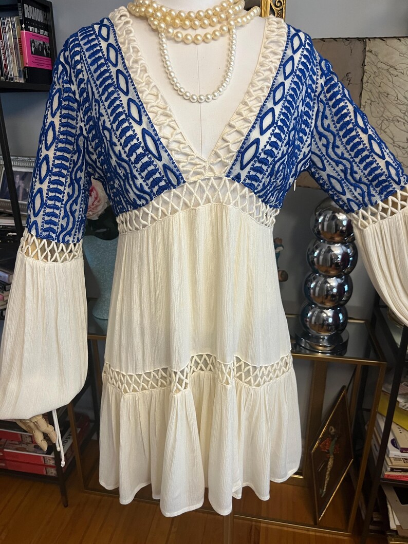 Pu&ograve; includere: Un abito color crema con maniche e corpetto ricamati in blu. L'abito presenta uno scollo a V, dettagli traforati e maniche lunghe a campana. Una collana di perle &egrave; drappeggiata intorno allo scollo. L'abito &egrave; esposto su un manichino.