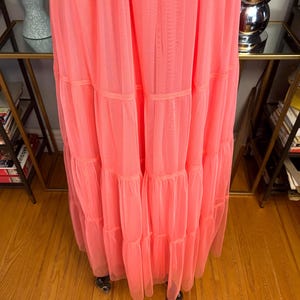 Puede incluir: Un vestido largo de color coral con m&uacute;ltiples volantes en capas. El vestido est&aacute; hecho de una tela transparente y ligera, y las capas est&aacute;n separadas por bandas del mismo material. El vestido se exhibe en un perchero rodante.