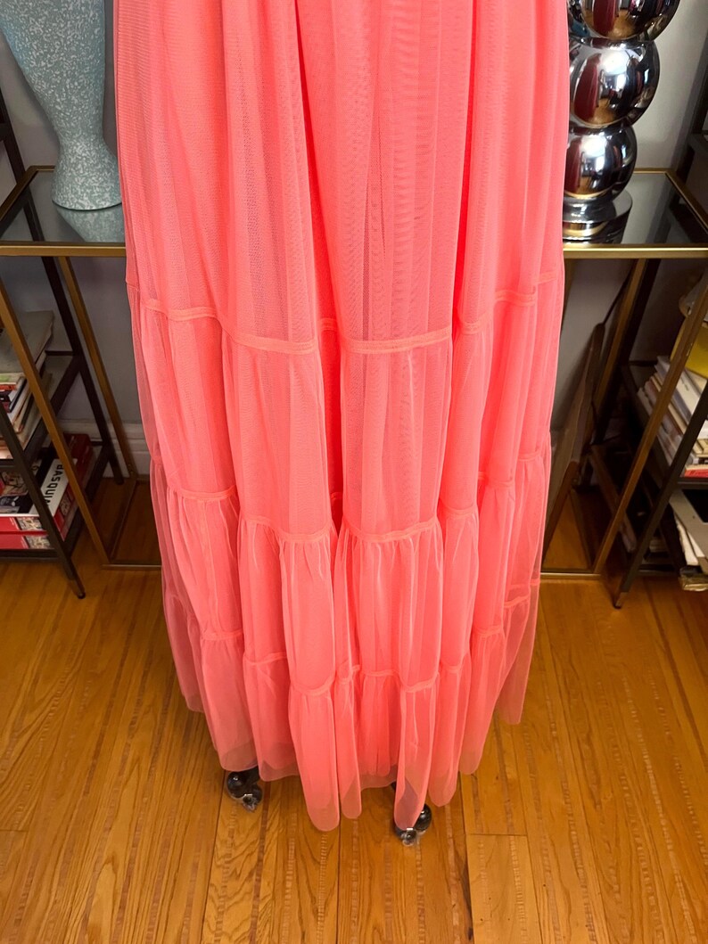 Puede incluir: Un vestido largo de color coral con m&uacute;ltiples volantes en capas. El vestido est&aacute; hecho de una tela transparente y ligera, y las capas est&aacute;n separadas por bandas del mismo material. El vestido se exhibe en un perchero rodante.