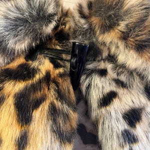 K&ouml;nnte beinhalten: Ein Kunstpelz-Kleidungsst&uuml;ck mit Leopardenmuster in Braun-, Schwarz- und Cremet&ouml;nen. Das Teil wird in der Mitte mit einem schwarzen Knebelverschluss aus glattem, hartem Material verschlossen, der an einer schwarzen Kordelschlaufe befestigt ist.
