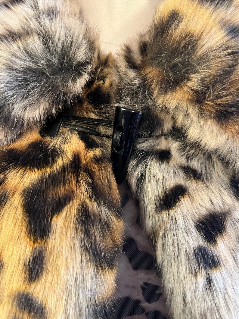 K&ouml;nnte beinhalten: Ein Kunstpelz-Kleidungsst&uuml;ck mit Leopardenmuster in Braun-, Schwarz- und Cremet&ouml;nen. Das Teil wird in der Mitte mit einem schwarzen Knebelverschluss aus glattem, hartem Material verschlossen, der an einer schwarzen Kordelschlaufe befestigt ist.