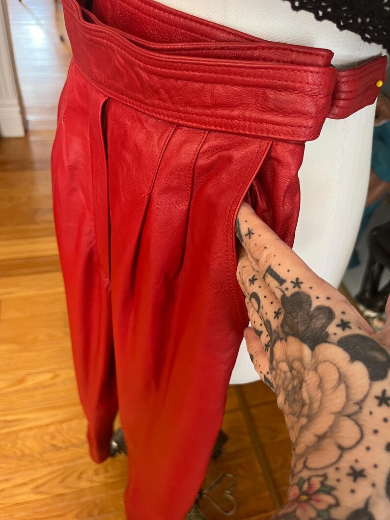 May include: Bright red leather pants with a high waist and pleated front. The pants feature a wide belt and pockets. The leather appears smooth and has a slight sheen. The pants are displayed on a mannequin.