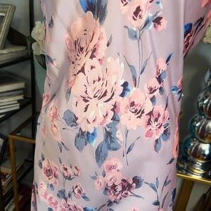 May include: A light purple fabric with a floral pattern featuring large pink and purple roses, blue accents, and green stems. The fabric has a smooth, silky texture and is likely used for clothing or home decor.