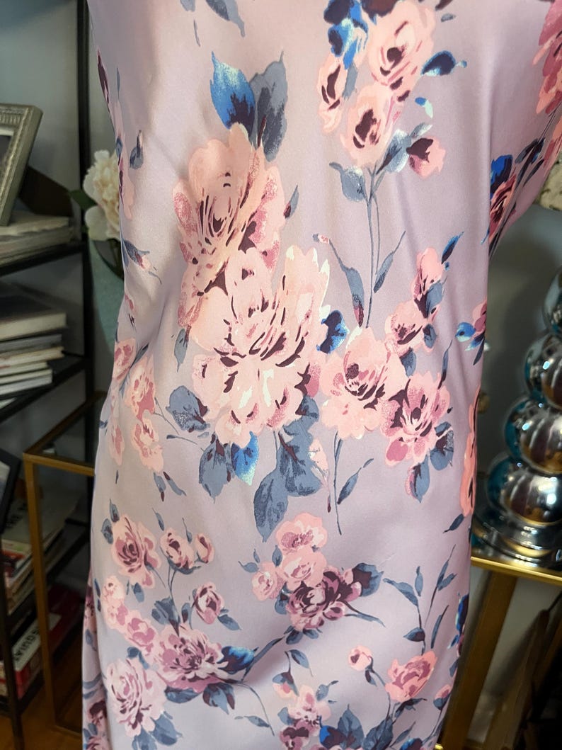 May include: A light purple fabric with a floral pattern featuring large pink and purple roses, blue accents, and green stems. The fabric has a smooth, silky texture and is likely used for clothing or home decor.