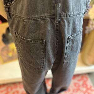 May include: Close-up of the back of a pair of charcoal-colored denim overalls. The overalls have two large back pockets and a small belt loop detail. The fabric appears to be a medium-weight denim.