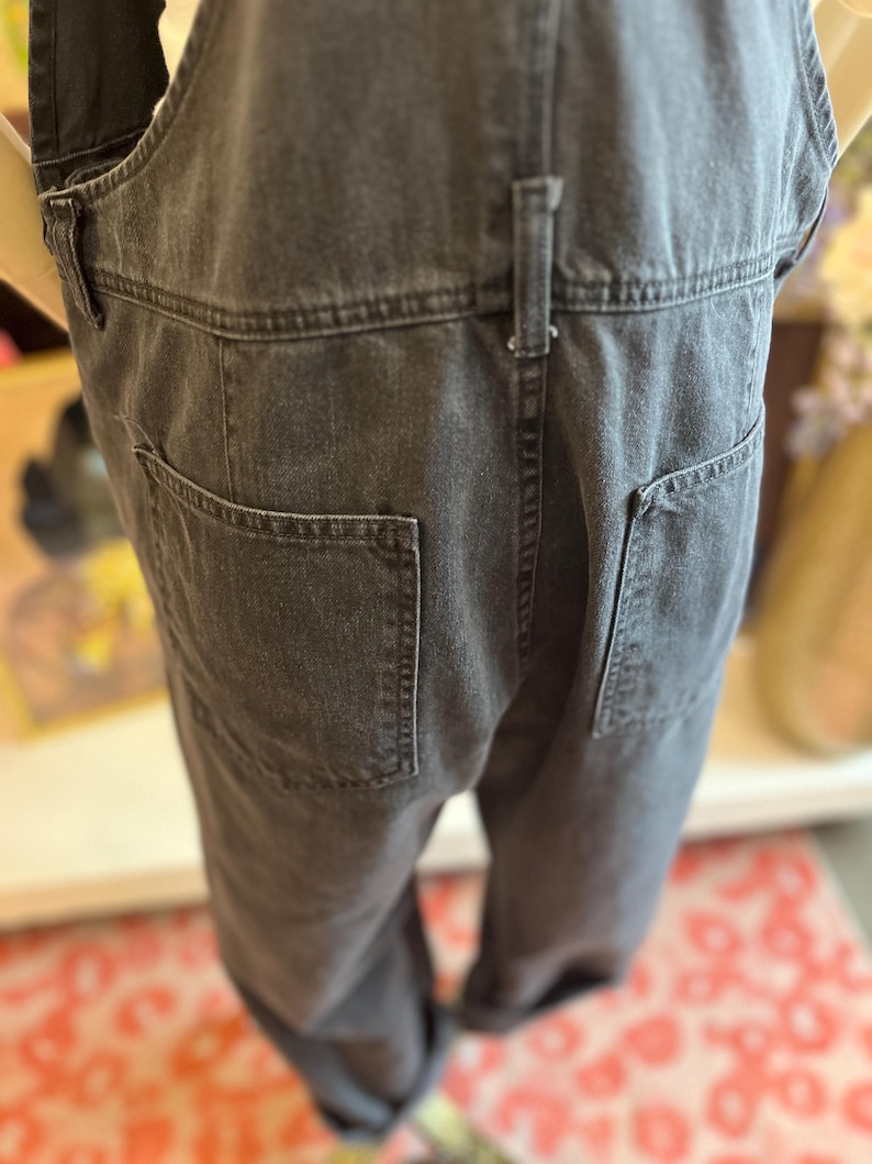 May include: Close-up of the back of a pair of charcoal-colored denim overalls. The overalls have two large back pockets and a small belt loop detail. The fabric appears to be a medium-weight denim.