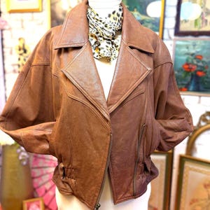 May include: A brown leather jacket featuring a wide collar, long sleeves, and a zippered front closure. The jacket is layered over a white top and accented with a leopard print scarf tied around the neck, showcasing a textured finish.