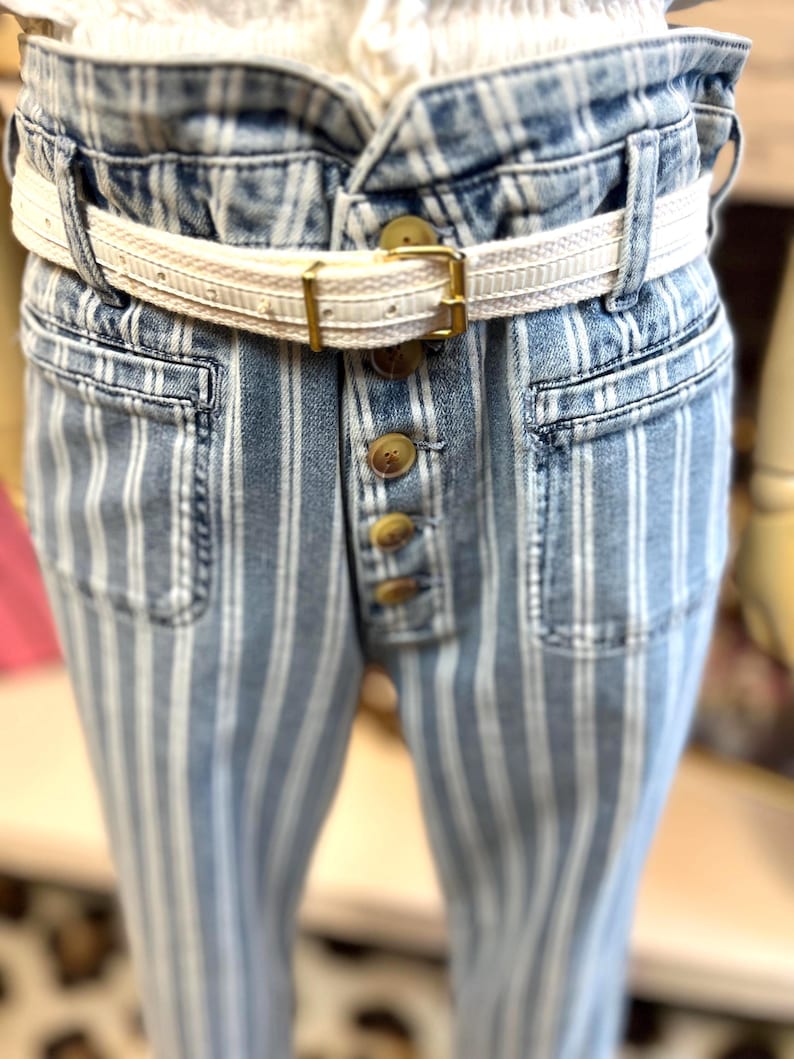 Pode incluir: Cal&ccedil;as de ganga azuis com riscas brancas verticais. As cal&ccedil;as de ganga t&ecirc;m cintura alta, braguilha com bot&otilde;es e cinto branco com fivela dourada. Os bolsos s&atilde;o vis&iacute;veis e o estilo geral &eacute; casual e moderno.