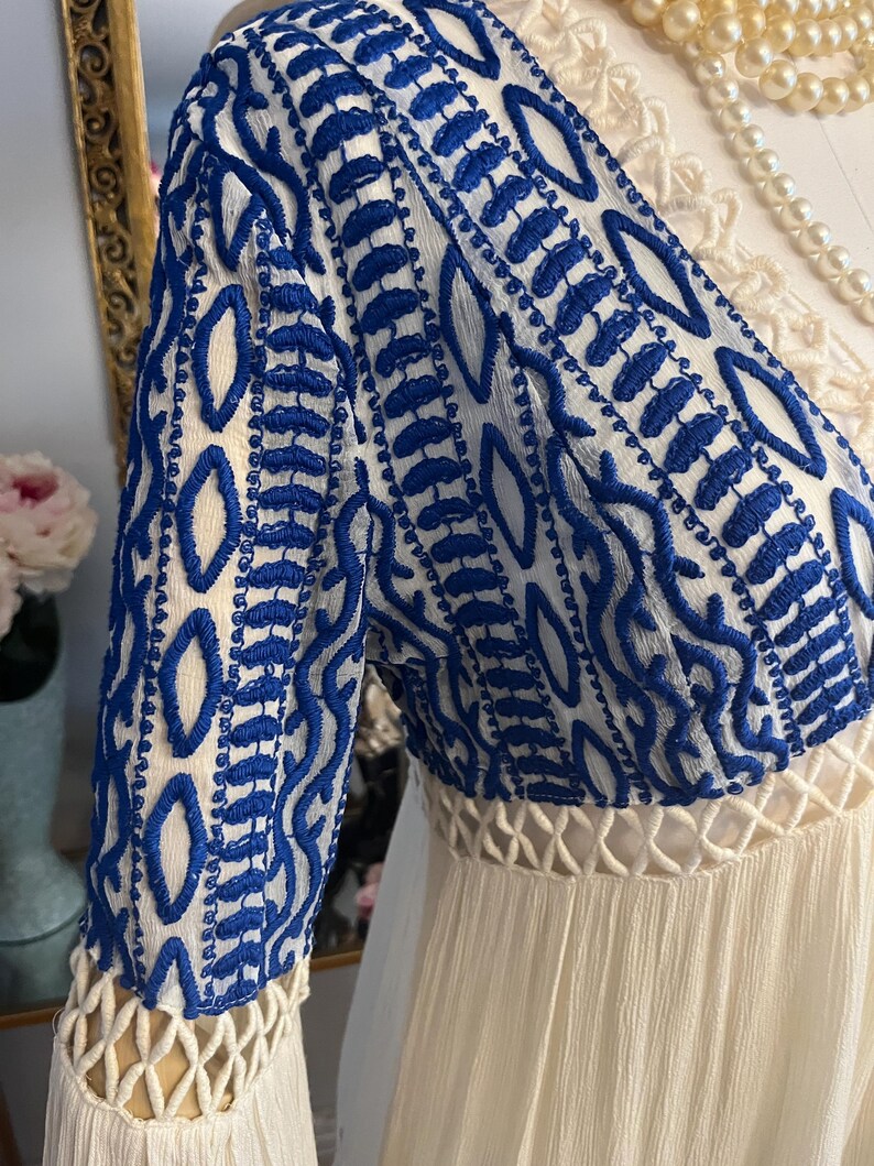 Pu&ograve; includere: Abito bianco con maniche ricamate blu. Le maniche presentano un motivo dettagliato di cuciture blu su sfondo bianco. L'abito ha una gonna fluida e strutturata e una vita decorativa.