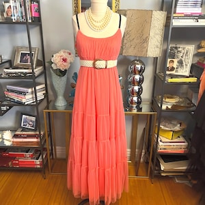 Puede incluir: Un vestido largo color coral con tirantes finos y falda escalonada. El vestido se ci&ntilde;e a la cintura con un cintur&oacute;n blanco tejido con hebilla dorada. El vestido se exhibe en un maniqu&iacute;, adornado con un collar de perlas.