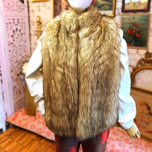 May include: A sleeveless vest featuring long, thick, light brown and tan faux fur panels that run vertically down the front. The vest is worn over a plain white long-sleeved shirt and dark patterned trousers, creating a layered appearance.