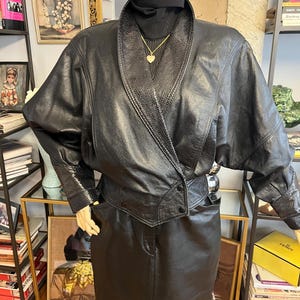 Puede incluir: Una chaqueta de cuero negro con un cuello chal ancho con un patrón texturizado similar a la piel de serpiente, mangas largas con puños texturizados a juego y un cierre frontal cruzado con dos botones visibles, sobre una prenda negra de cuello alto.