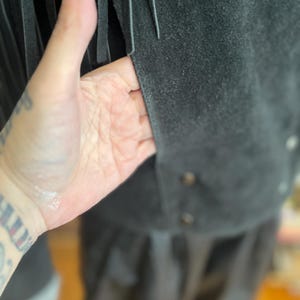 May include: Close-up of a dark gray suede jacket with long fringe detailing. The jacket has a button closure and a soft, textured appearance. The fringe hangs vertically, adding a decorative element to the garment.