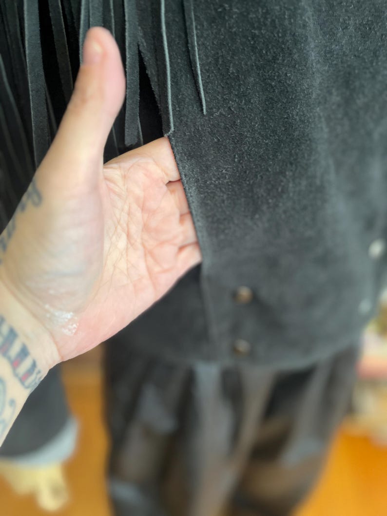 May include: Close-up of a dark gray suede jacket with long fringe detailing. The jacket has a button closure and a soft, textured appearance. The fringe hangs vertically, adding a decorative element to the garment.