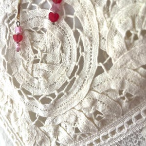 Op de afbeelding: Close-up van delicate, cr&egrave;mekleurige kanten stof met een bloemmotief. Hangende oorbellen met goudkleurige staven en rode en roze hartvormige kralen zijn zichtbaar. De oorbellen voegen een vleugje kleur en contrast toe aan de zachte, getextureerde achtergrond.