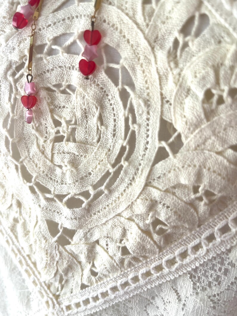 Op de afbeelding: Close-up van delicate, cr&egrave;mekleurige kanten stof met een bloemmotief. Hangende oorbellen met goudkleurige staven en rode en roze hartvormige kralen zijn zichtbaar. De oorbellen voegen een vleugje kleur en contrast toe aan de zachte, getextureerde achtergrond.