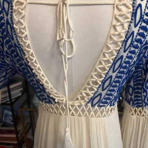 Pu&ograve; includere: Abito bianco con dettagli ricamati blu e schiena scoperta. L'abito presenta un bordo color crema a trama aperta lungo lo scollo e le maniche. Un nappa color crema pende dalla schiena dell'abito.