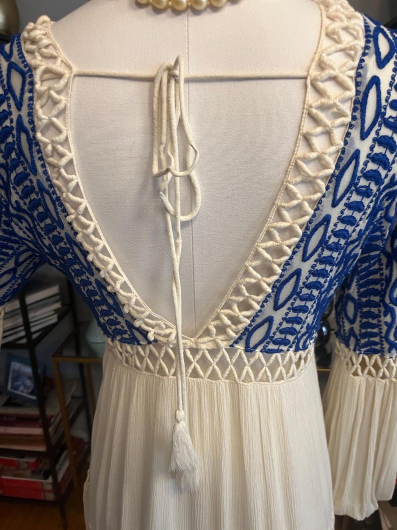 Pu&ograve; includere: Abito bianco con dettagli ricamati blu e schiena scoperta. L'abito presenta un bordo color crema a trama aperta lungo lo scollo e le maniche. Un nappa color crema pende dalla schiena dell'abito.