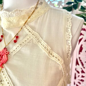 Op de afbeelding: Cr&egrave;mekleurige mouwloze top met kanten rand en een hoge halslijn. De top heeft delicate kanten details langs de randen en een decoratief kanten paneel over de borst. Een gouden ketting met rode hartvormige hangers hangt ervoor.