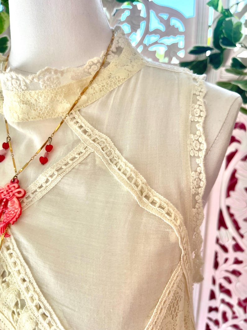 Op de afbeelding: Cr&egrave;mekleurige mouwloze top met kanten rand en een hoge halslijn. De top heeft delicate kanten details langs de randen en een decoratief kanten paneel over de borst. Een gouden ketting met rode hartvormige hangers hangt ervoor.