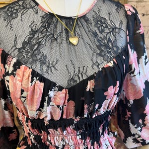 May include: A black floral print blouse with pink flowers and a black lace neckline. The blouse has long sleeves and a fitted waist. A gold heart-shaped locket hangs from a delicate gold chain.