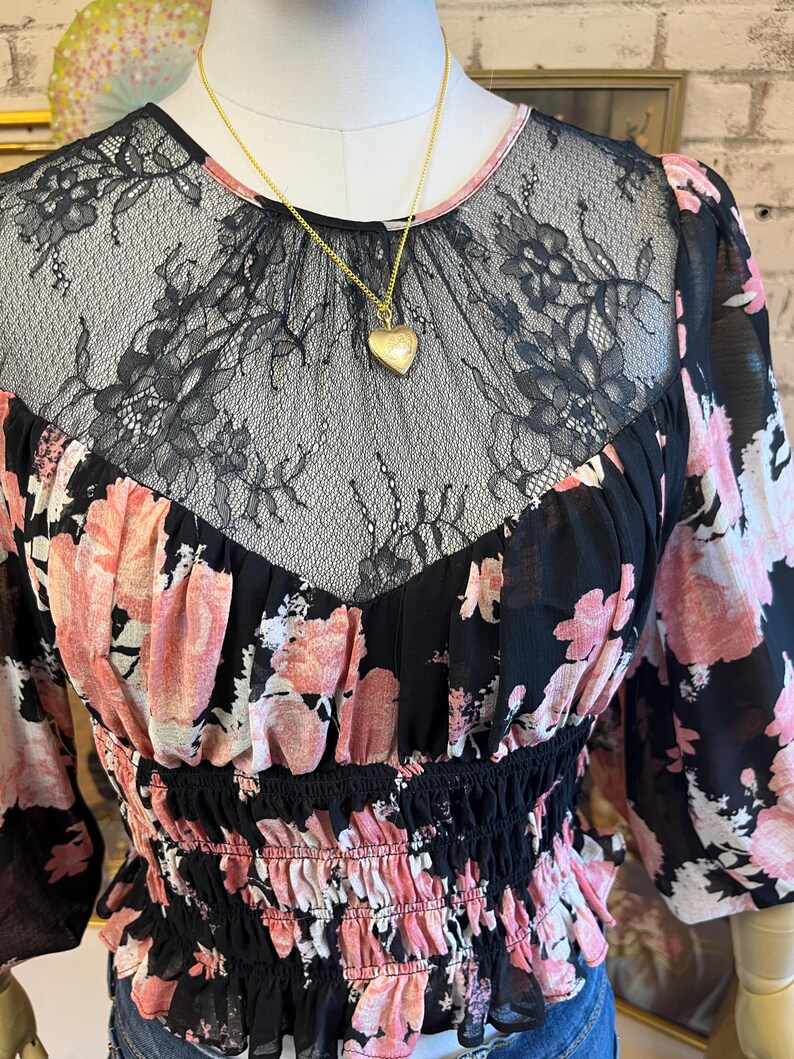 May include: A black floral print blouse with pink flowers and a black lace neckline. The blouse has long sleeves and a fitted waist. A gold heart-shaped locket hangs from a delicate gold chain.