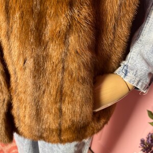 May include: A brown fur coat with a textured appearance. The coat is worn over a light blue denim jacket and light blue jeans. The fur appears to be a warm, reddish-brown color.