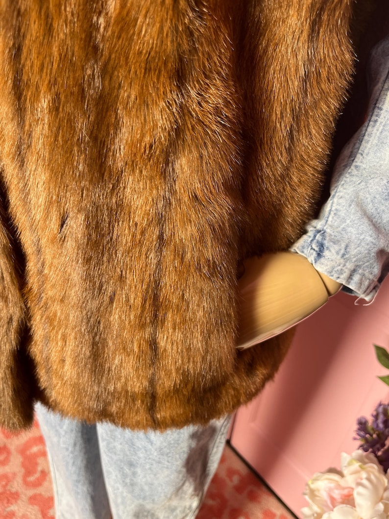 May include: A brown fur coat with a textured appearance. The coat is worn over a light blue denim jacket and light blue jeans. The fur appears to be a warm, reddish-brown color.