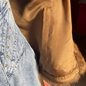 May include: Close-up of a tan-colored coat sleeve with a brown fur trim. The coat's interior lining is a smooth, shiny fabric. A denim jacket with gold-colored rivets is visible on the left side of the image.