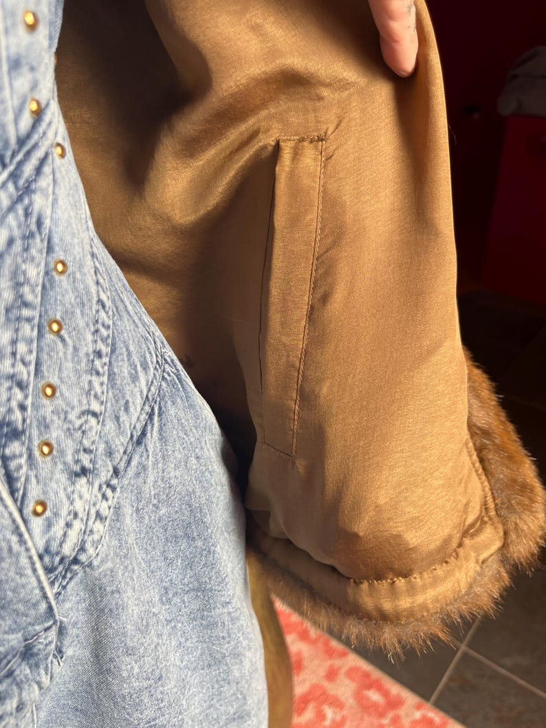 May include: Close-up of a tan-colored coat sleeve with a brown fur trim. The coat's interior lining is a smooth, shiny fabric. A denim jacket with gold-colored rivets is visible on the left side of the image.