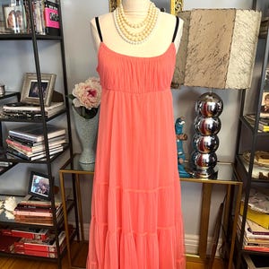 Puede incluir: Un vestido largo color coral con tirantes finos y falda escalonada se exhibe en un maniqu&iacute;. El vestido tiene un corpi&ntilde;o fruncido y una tela fluida y ligera. Un collar de perlas de varias hebras est&aacute; drapeado alrededor del cuello del maniqu&iacute;.