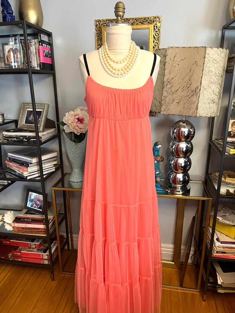Puede incluir: Un vestido largo color coral con tirantes finos y falda escalonada se exhibe en un maniqu&iacute;. El vestido tiene un corpi&ntilde;o fruncido y una tela fluida y ligera. Un collar de perlas de varias hebras est&aacute; drapeado alrededor del cuello del maniqu&iacute;.