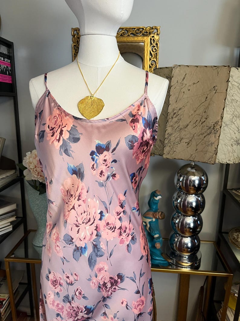 May include: A light purple slip dress with a floral pattern in shades of pink, blue, and purple. The dress has thin straps and is displayed on a mannequin. A gold leaf pendant hangs from a delicate chain.