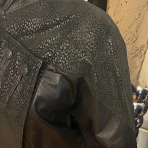 May include: Black leather jacket with a textured, spotted pattern on the shoulders and upper sleeves. The jacket features a high collar, button details, and a contrasting smooth leather panel. The overall design is a mix of textures and shapes.