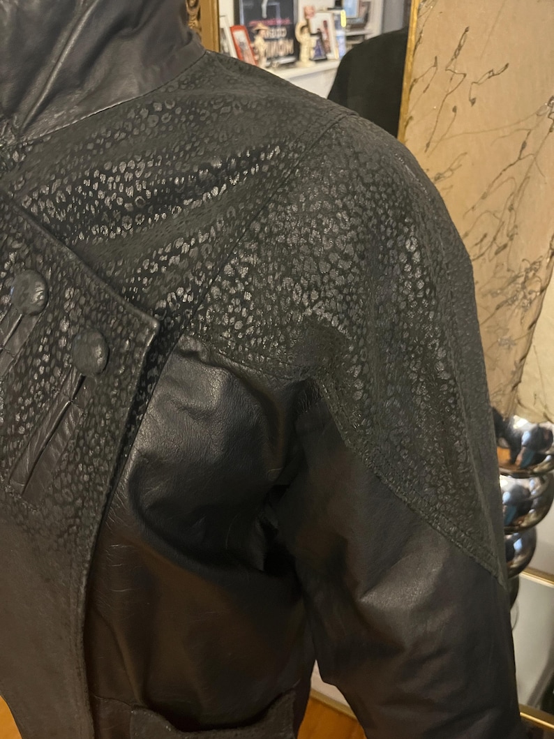 May include: Black leather jacket with a textured, spotted pattern on the shoulders and upper sleeves. The jacket features a high collar, button details, and a contrasting smooth leather panel. The overall design is a mix of textures and shapes.