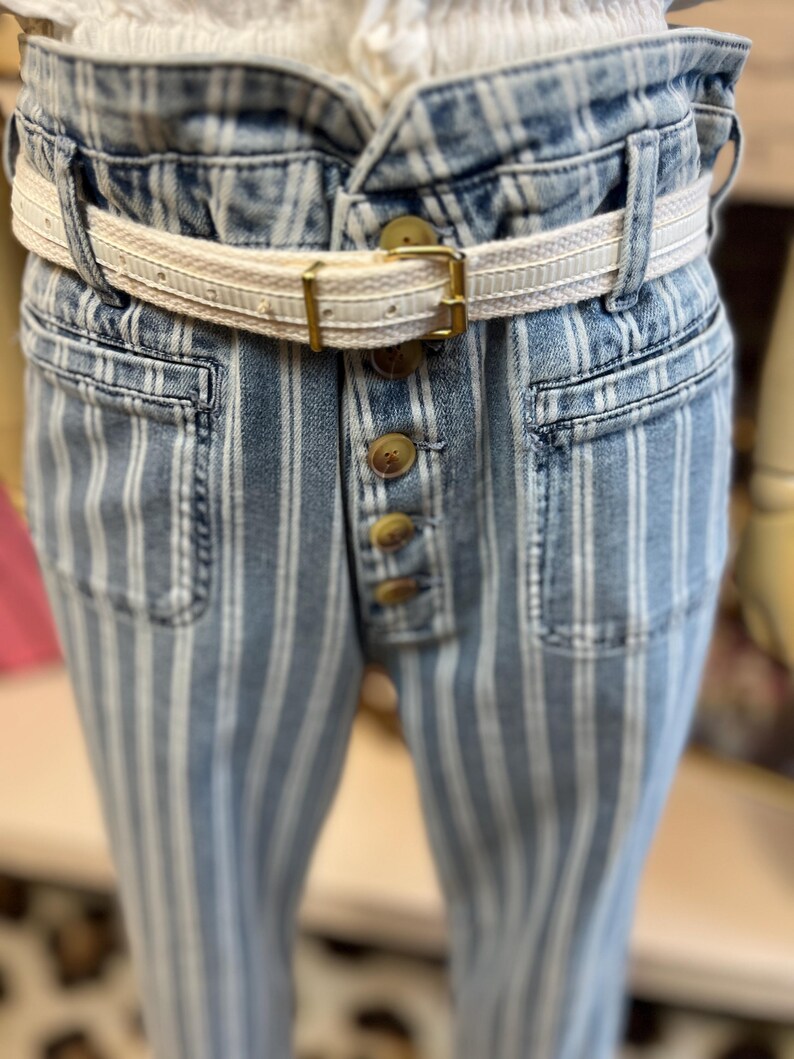 Pode incluir: Jeans azuis e brancos listrados com cintura alta e braguilha de bot&otilde;es. Os jeans t&ecirc;m um cinto creme com fivela dourada. Os bolsos s&atilde;o vis&iacute;veis e o tecido tem um aspeto ligeiramente desbotado.