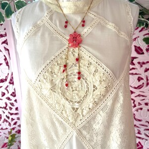 Op de afbeelding: Een cr&egrave;mekleurige mouwloze jurk met kanten details en een hoge halslijn. Een goudkleurige ketting met een roze hanger en rode hartvormige kralen hangt aan de jurk. De jurk heeft een vintage uitstraling.