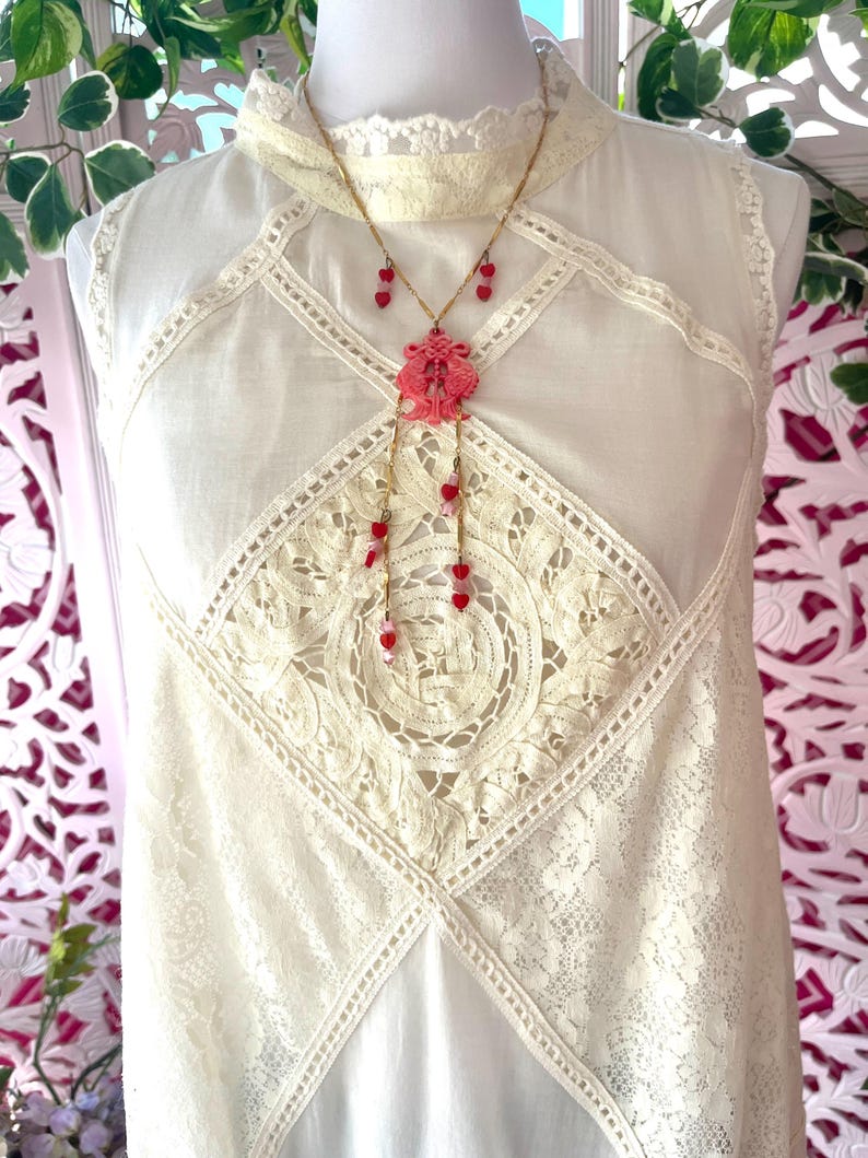 Op de afbeelding: Een cr&egrave;mekleurige mouwloze jurk met kanten details en een hoge halslijn. Een goudkleurige ketting met een roze hanger en rode hartvormige kralen hangt aan de jurk. De jurk heeft een vintage uitstraling.