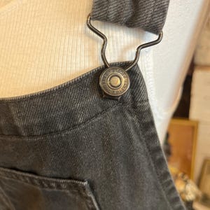 May include: Close-up of a dark gray denim overall. The overall features a metal clasp and a button with the text "FREE PEOPLE" around the edge. The overall has a pocket and a strap.