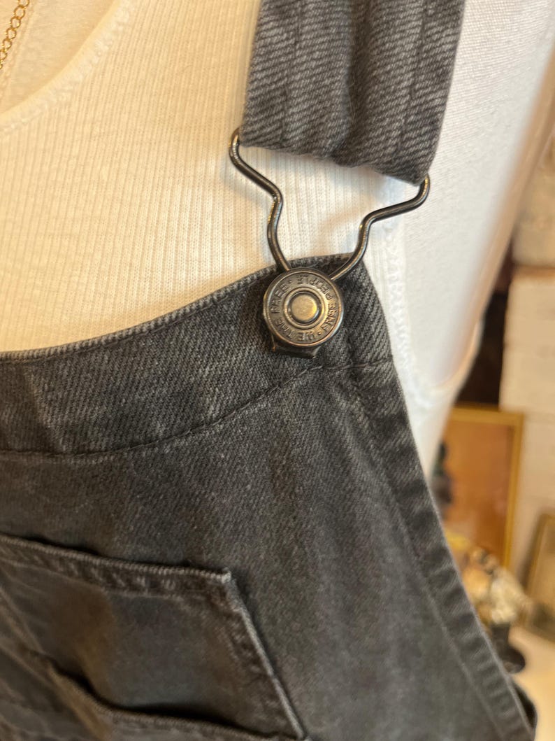 May include: Close-up of a dark gray denim overall. The overall features a metal clasp and a button with the text "FREE PEOPLE" around the edge. The overall has a pocket and a strap.