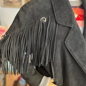 May include: Black suede jacket with fringe detail on the shoulders. The jacket features a classic collar, a button closure, and a decorative silver button. The sleeves have a gray knit cuff. The jacket is displayed on a mannequin.