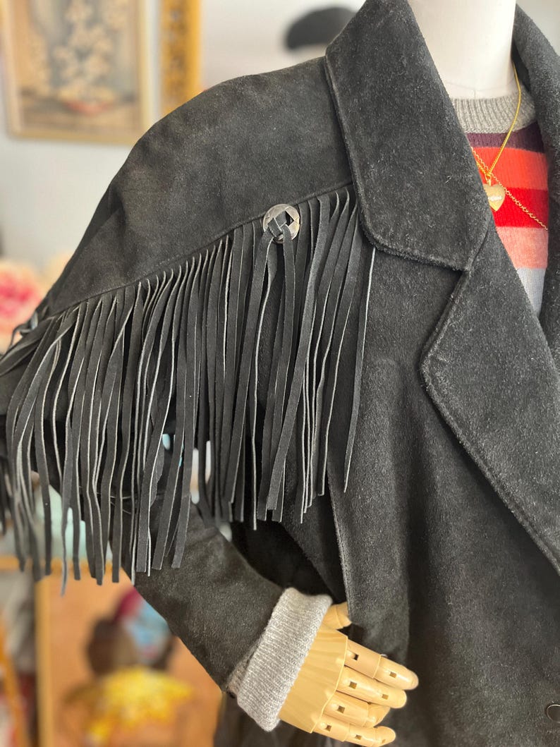May include: Black suede jacket with fringe detail on the shoulders. The jacket features a classic collar, a button closure, and a decorative silver button. The sleeves have a gray knit cuff. The jacket is displayed on a mannequin.
